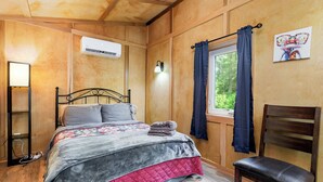 Une pièce aux murs lambrissés, comprenant un lit, une chaise, une fenêtre avec des rideaux, un appareil de climatisation et un luminaire fixé au mur.