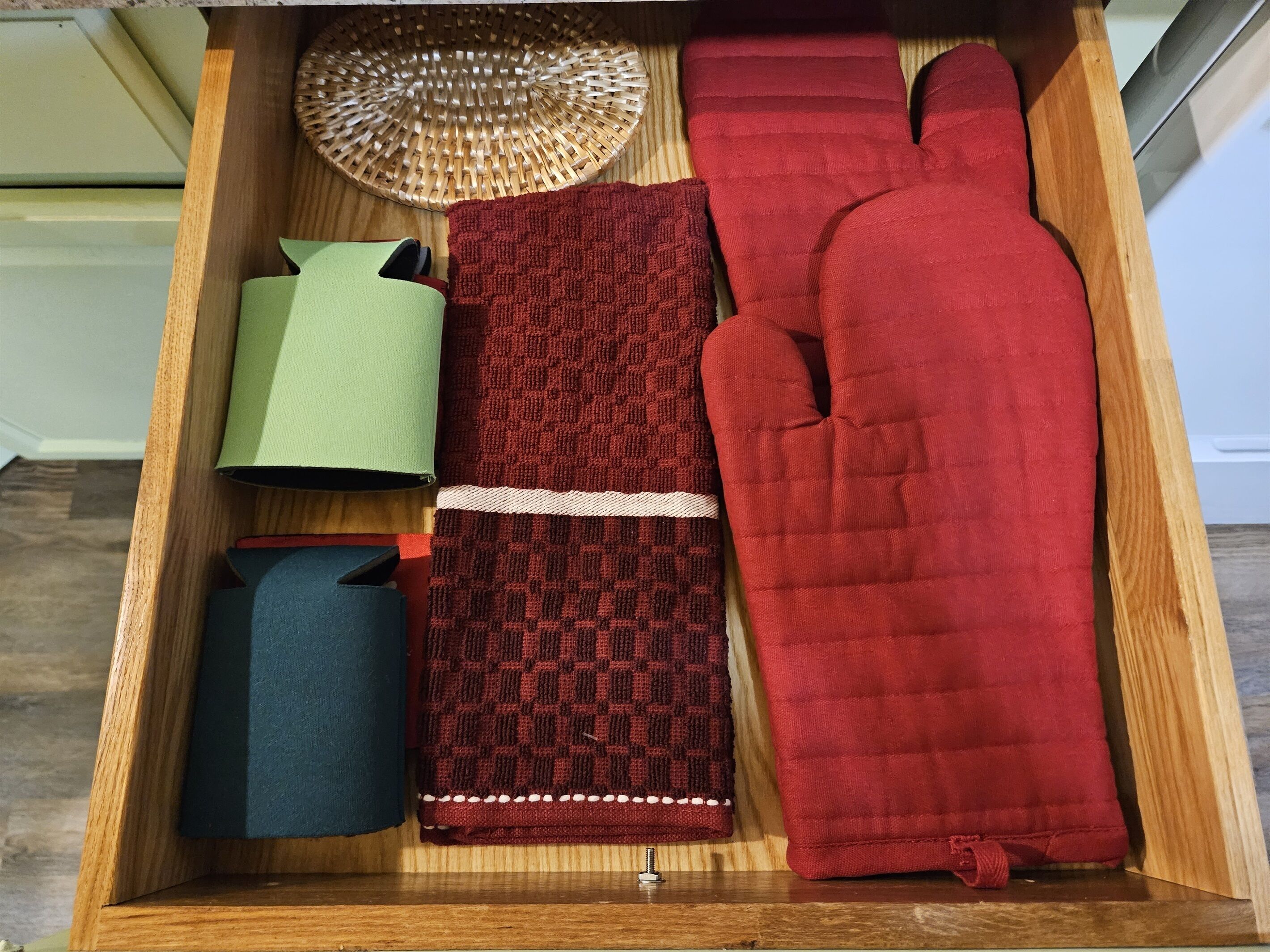 Een houten lade met diverse keukenbenodigdheden, waaronder een rieten mand, een rode ovenhandschoen, een groene pan, een blauwe pan en een rood geruit stukje stof.