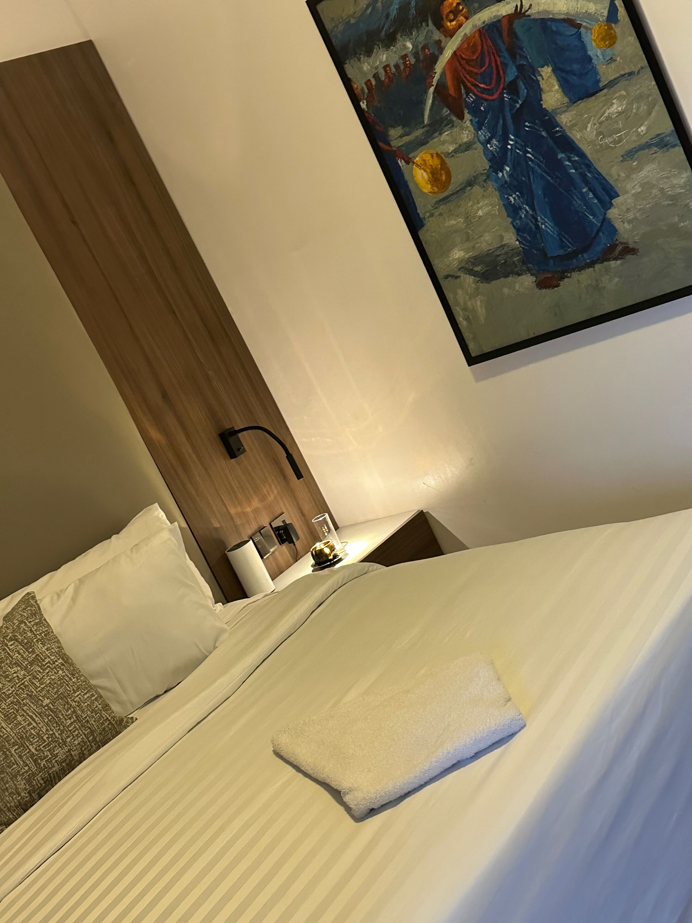 Ett hotellrum med en säng, kuddar, ett sängbord med en lampa och en tavla på väggen.
