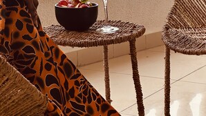 Un balcón con una silla de mimbre, una mesita con una bebida y fruta, y vistas de edificios a través de un cuerpo de agua.