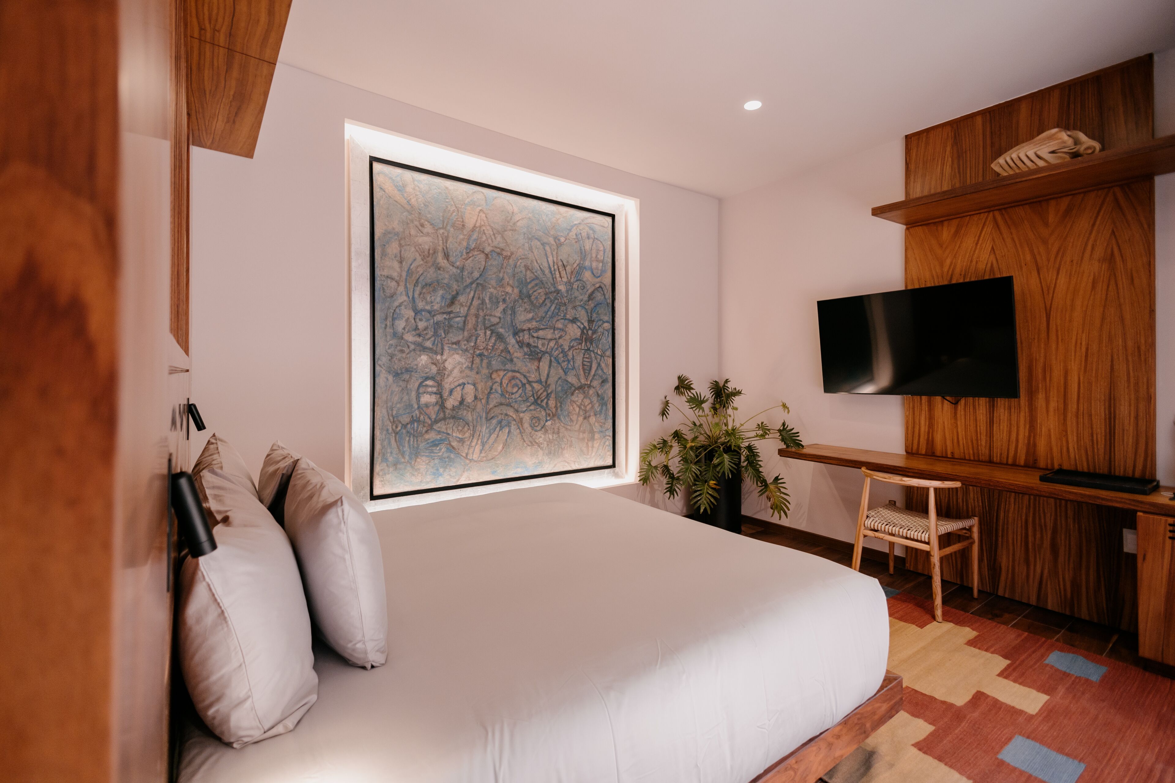 A modern bedroom with a large bed, a mounted TV, a wooden desk, and a large abstract painting.