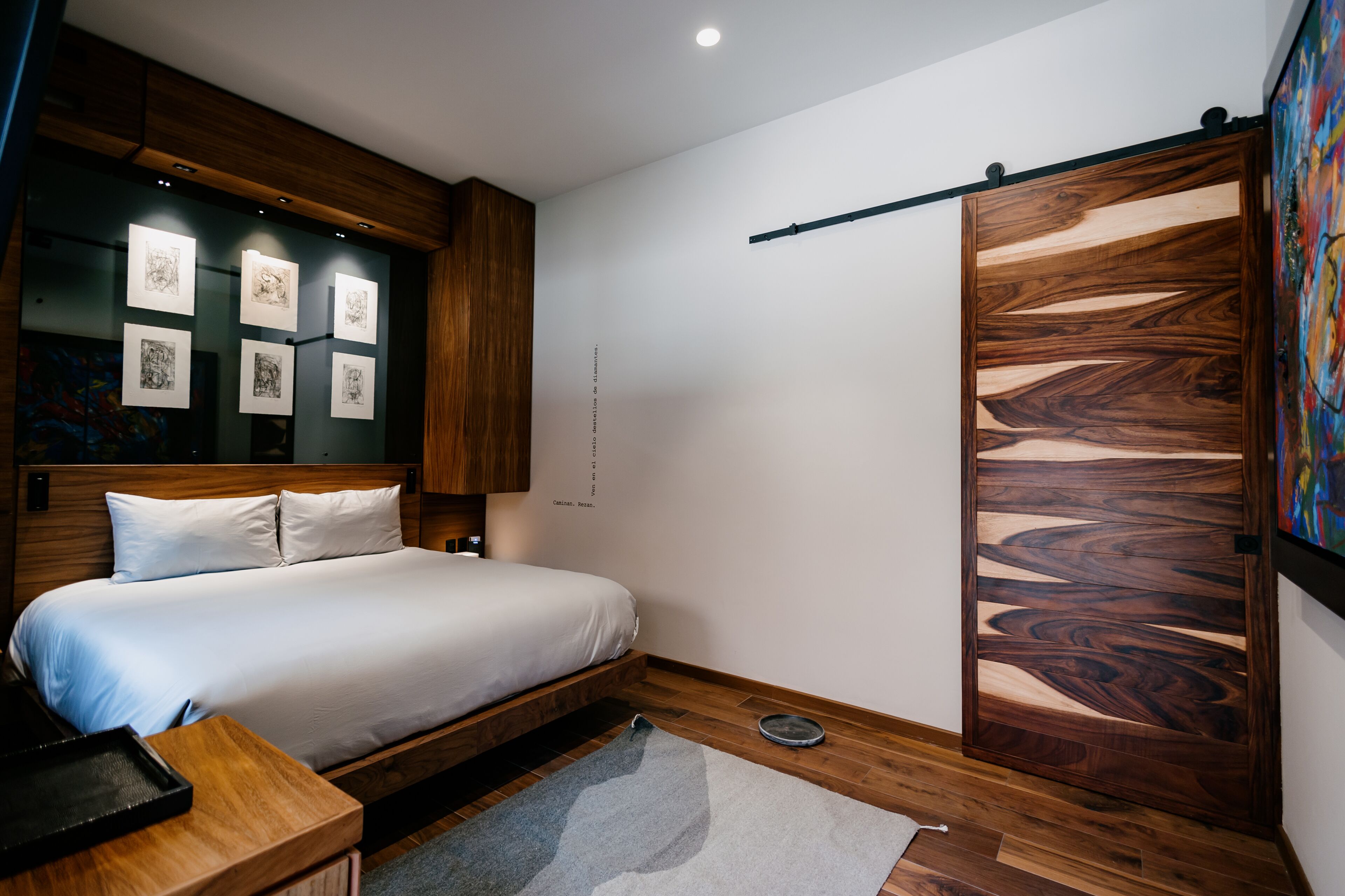 A modern bedroom with a wooden headboard, a bed with white bedding, a bedside table, a wall-mounted shelf with artwork, and a sliding wooden door.