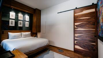 A modern bedroom with a wooden headboard, a bed with white bedding, a bedside table, a wall-mounted shelf with artwork, and a sliding wooden door.