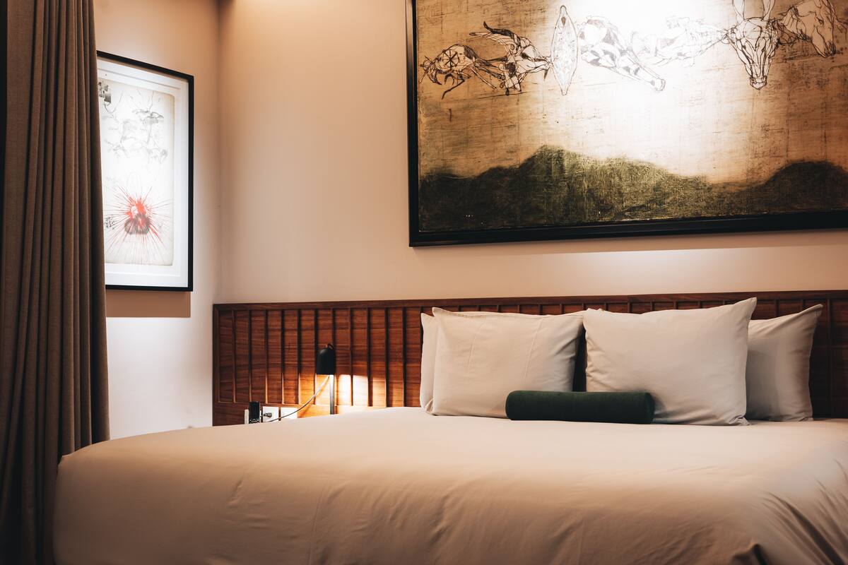 A neatly made bed with white linens, a wooden headboard, and a framed artwork above it.
