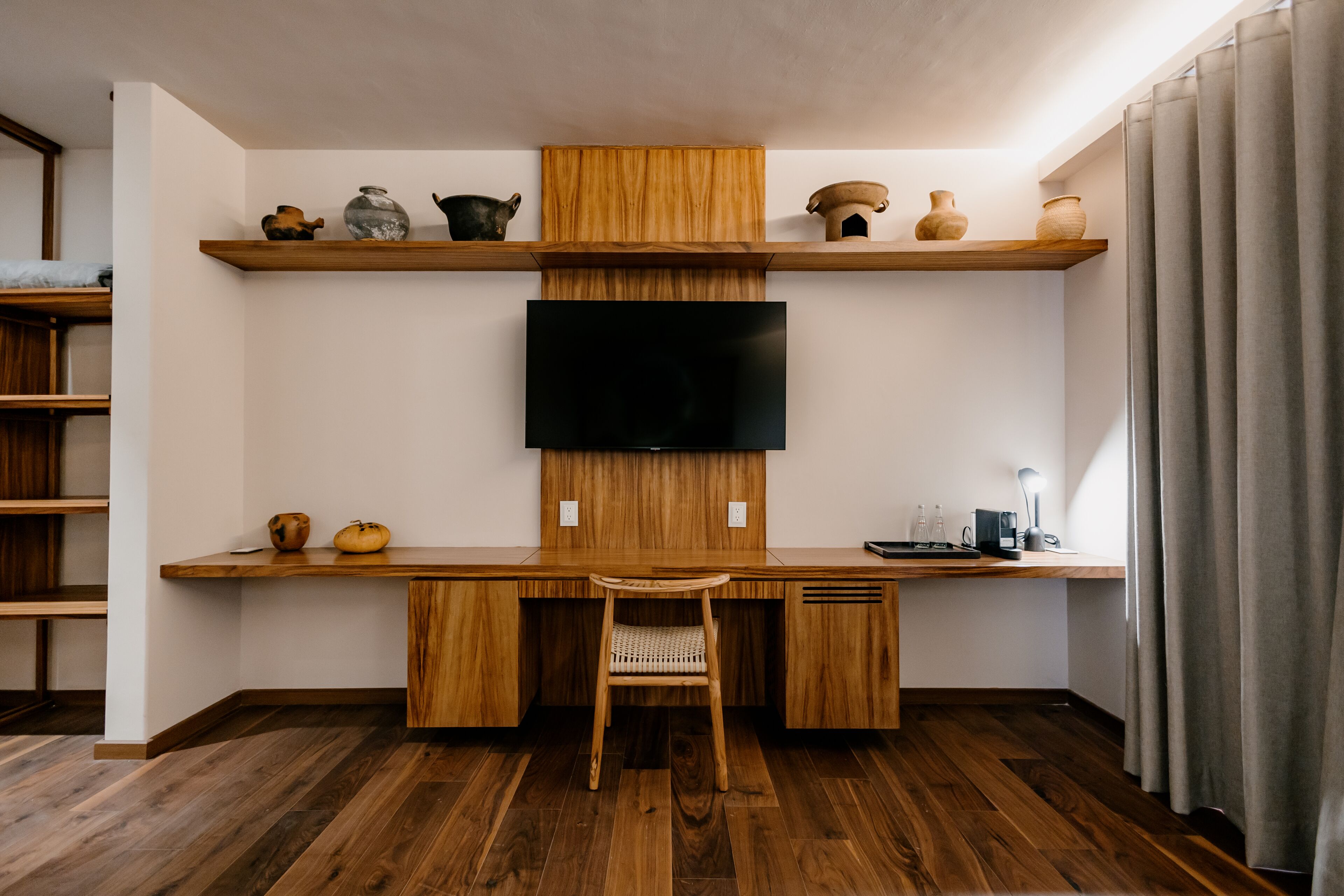 Une chambre moderne avec un bureau en bois, un téléviseur fixé au mur et des étagères ornées d’objets décoratifs.