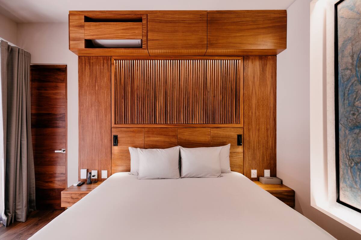 A modern hotel room with a wooden headboard, a large bed, bedside tables, and a window.