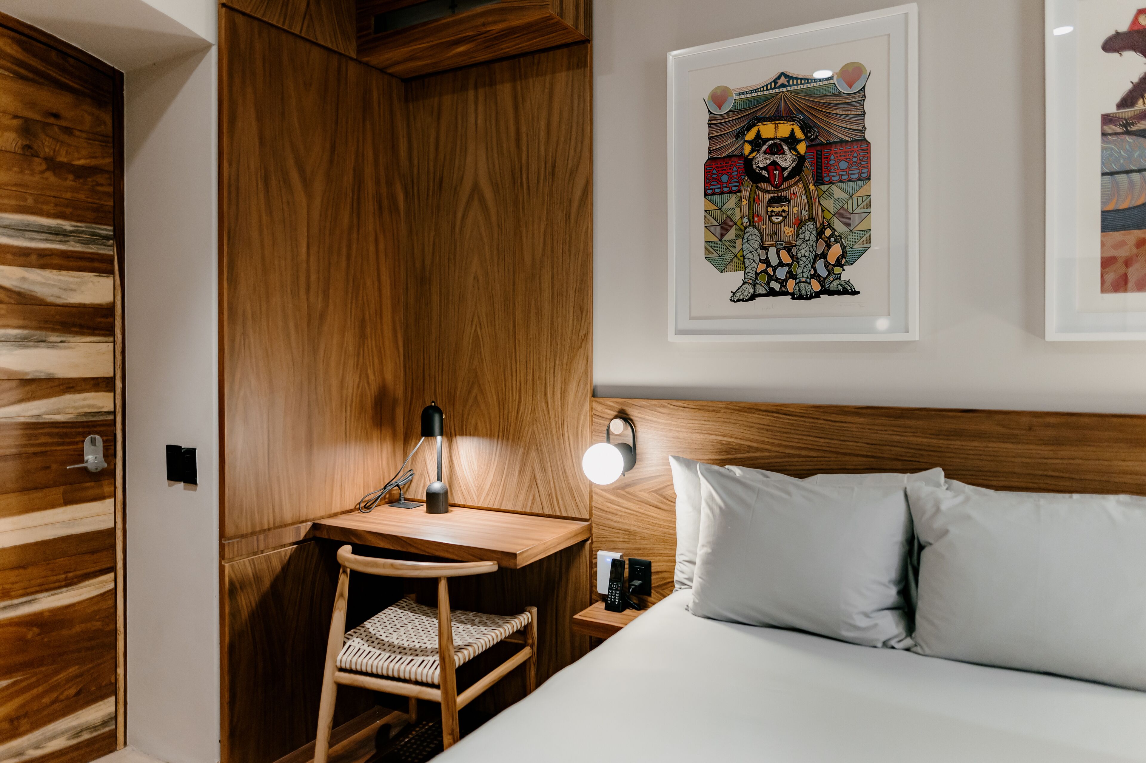 Habitación de hotel con un armario de madera, un escritorio con una silla, una mesita de noche con una lámpara y una cama con almohadas.