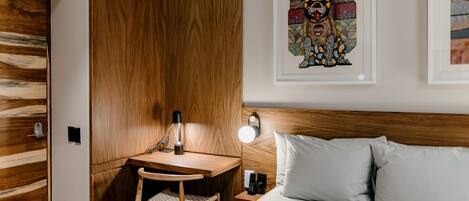 Habitación de hotel con un armario de madera, un escritorio con una silla, una mesita de noche con una lámpara y una cama con almohadas.