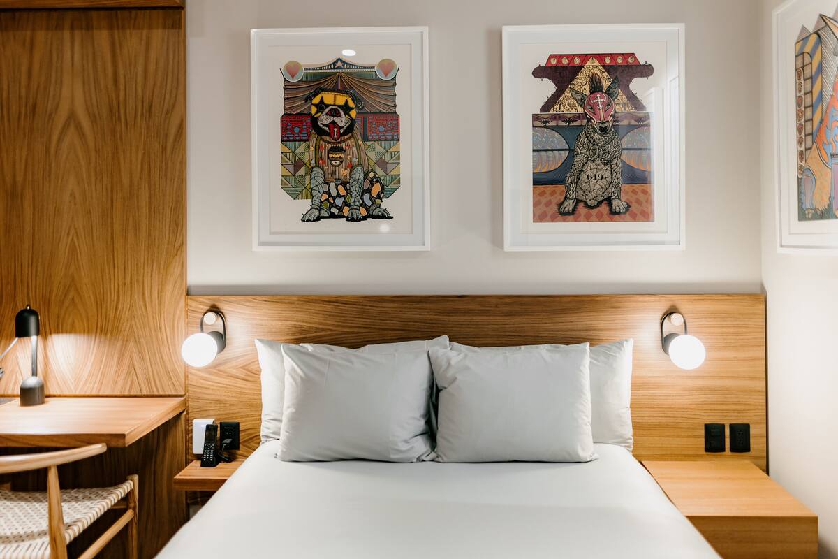 A modern hotel room with a wooden headboard, two bedside lamps, a bed with white linens, a wooden nightstand, and two framed artworks on the wall.