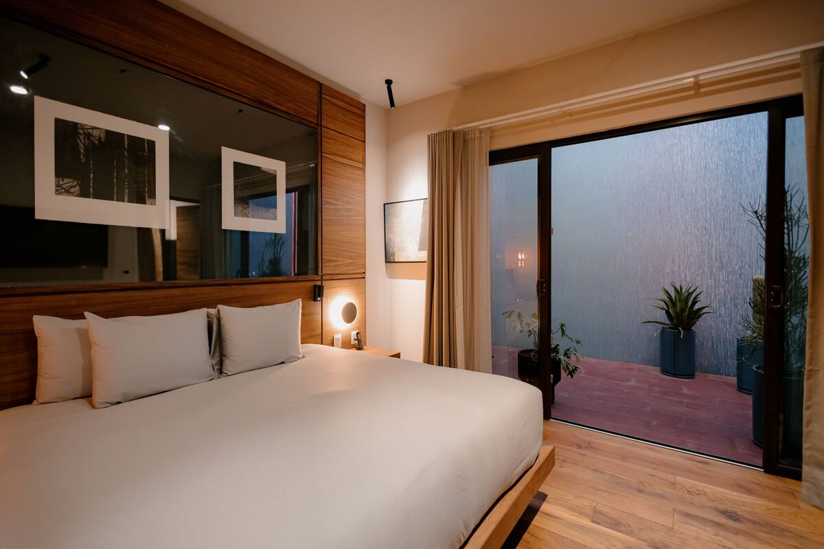 A modern hotel room with a large bed, wooden headboard, and a balcony with potted plants.