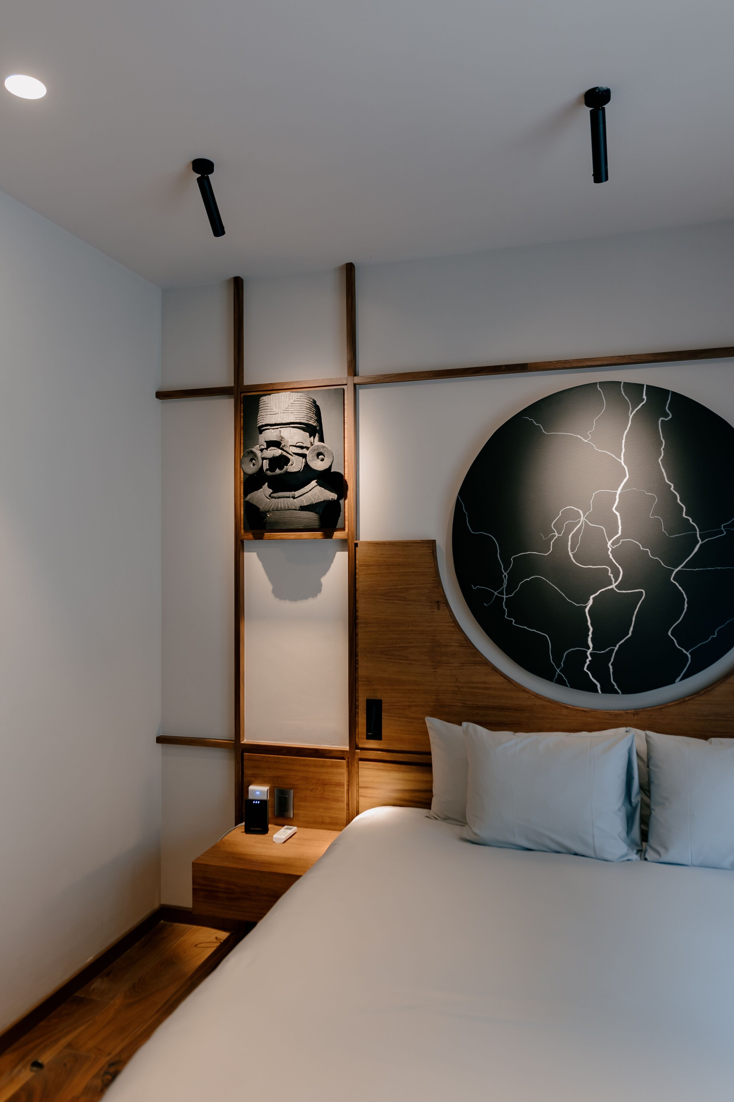 Ein modernes Hotelzimmer mit einem hölzernen Kopfteil, einem großen, runden Wandbild, einer kleinen Ablage mit einem dekorativen Gegenstand und einem Nachttisch mit Fernbedienung.