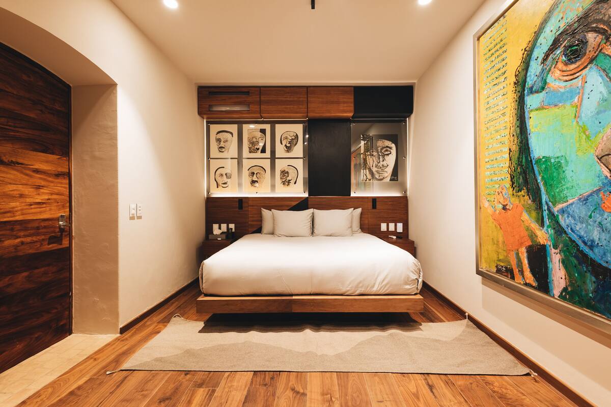 A bedroom with a bed, wooden headboard, artwork on the wall, and a large painting.