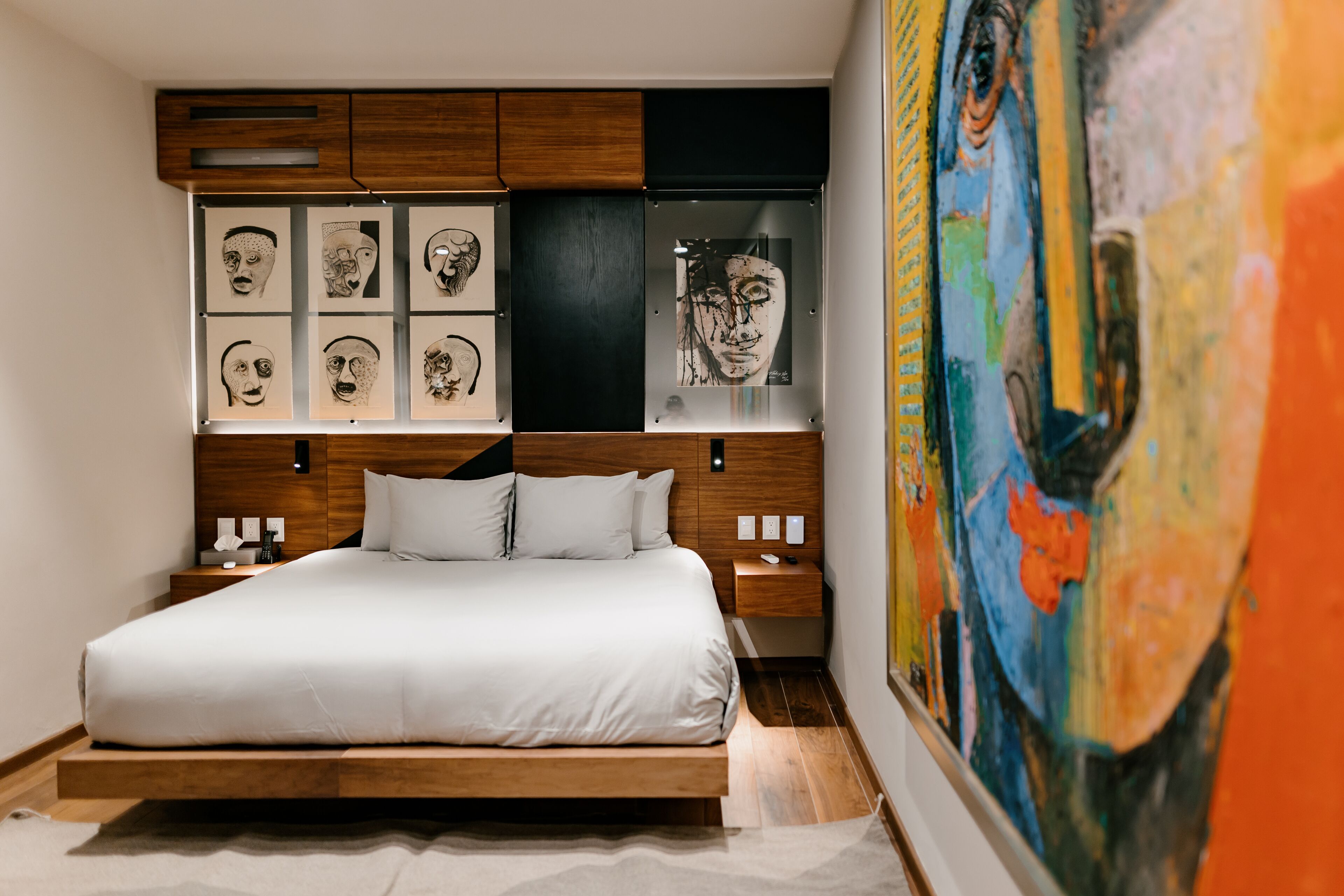 A modern bedroom with a wooden bed frame, white bedding, and a large abstract painting on the wall.