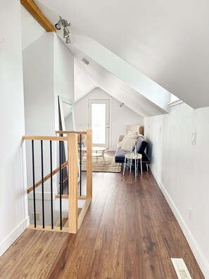 A spacious loft bedroom with a sloped ceiling, a wooden staircase, a large window, and a cozy seating area with a sofa and a side table. - The Doll House (Chilliwack)