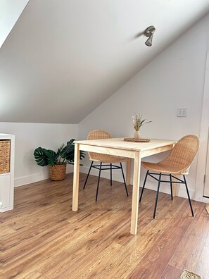 A dining area with a wooden table and two wicker chairs, a potted plant, and a ceiling light fixture. - The Doll House (Chilliwack)