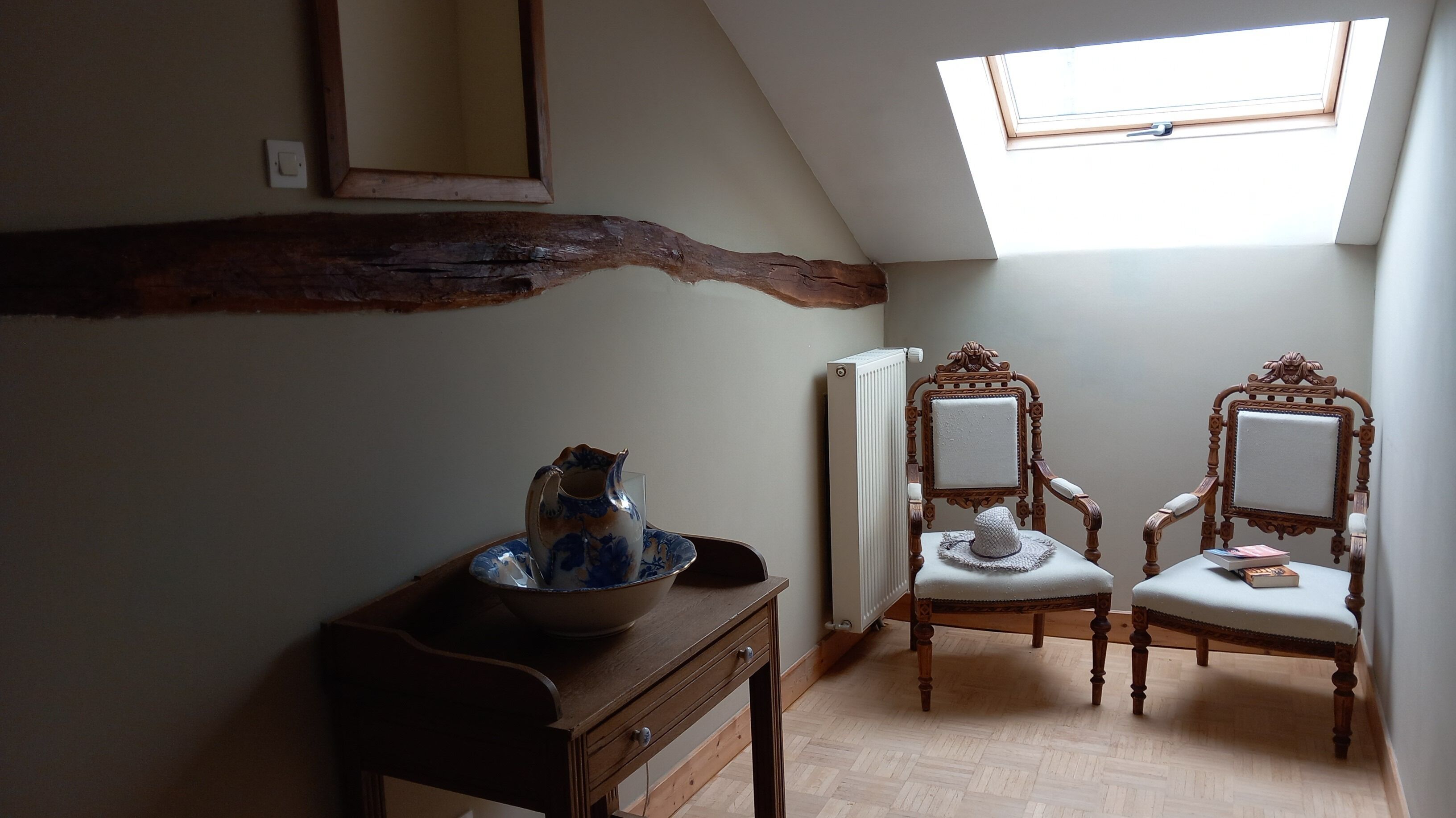 A room with a sloped ceiling, a skylight, a wooden beam, two ornate chairs, a table with a vase, and a radiator.