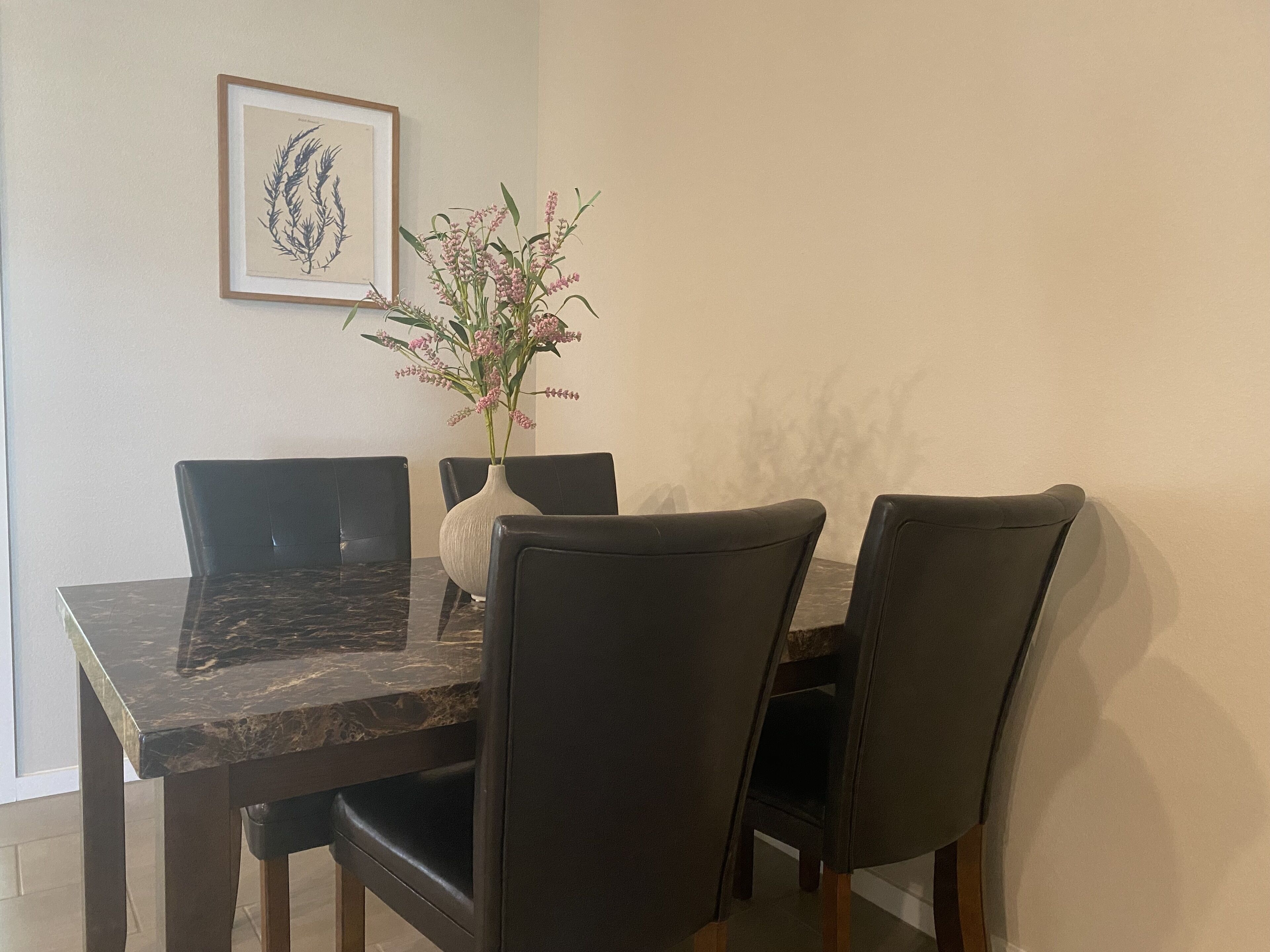 A dining table with a marble top, surrounded by four black chairs. A white vase with pink flowers is placed on the table.