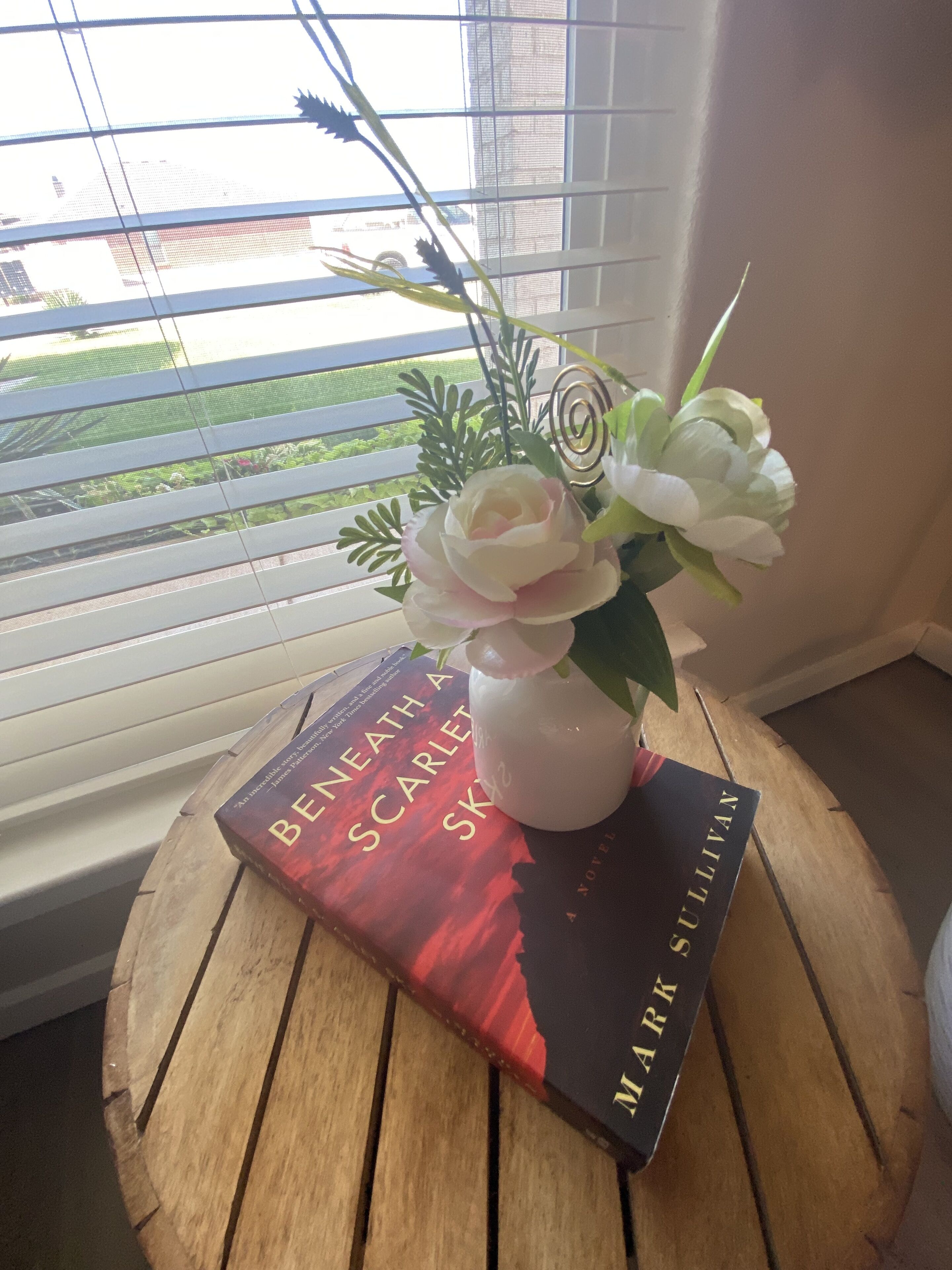 A book titled 'Beneath A Scarlett Sky' by Mark Sullivan on a wooden table near a window with blinds.