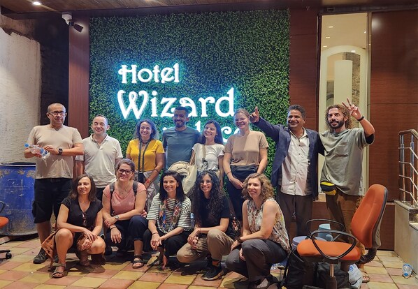 Um grupo de pessoas posando em frente a um letreiro do 'Hotel Wizard', com um fundo de folhagem verde.