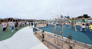 A playground with various play structures, a wooden fence, and a paved walkway. - Joyful Home near Monterey, Carmel, Pebble Beach, Big Sur- STR25-000023 (Marina)