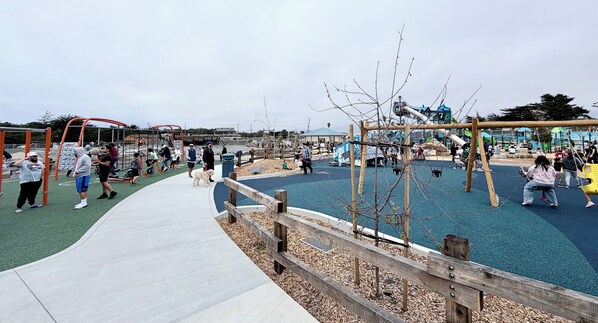 A playground with various play structures, a wooden fence, and a paved walkway. - Joyful Home near Monterey, Carmel, Pebble Beach, Big Sur- STR25-000023 (Marina)