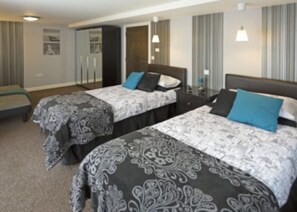 Two single beds with patterned bedspreads and pillows, a bench, and a table with a lamp.