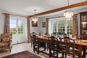 Une salle à manger avec une table en bois, des chaises et des lustres. On y trouve des fenêtres avec des rideaux, une porte et un tapis sur le sol.