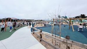 A playground with various play structures, a wooden fence, and a paved walkway. - The Ultimate Gateway, near Monterey, Carmel, Pebble Beach, Big Sur- STR25-000022 (Marina)