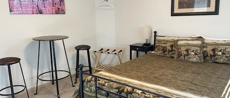 Un dormitorio con una cama, dos taburetes, una mesita, un perchero y dos cuadros en las paredes.