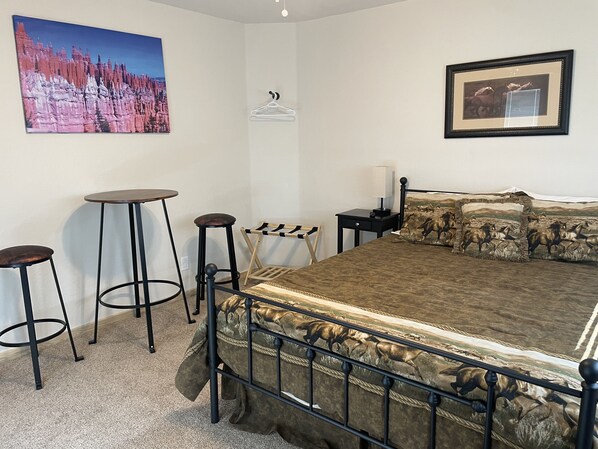 Un dormitorio con una cama, dos taburetes, una mesita, un perchero y dos cuadros en las paredes.