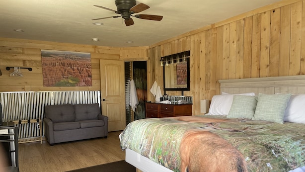 A bedroom with a large bed, a gray sofa, a wooden ceiling fan, and a painting of a landscape.