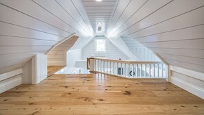 Een ruime zolderkamer met houten vloeren, witte wanden en een houten trap naar een hoger gelegen gedeelte.