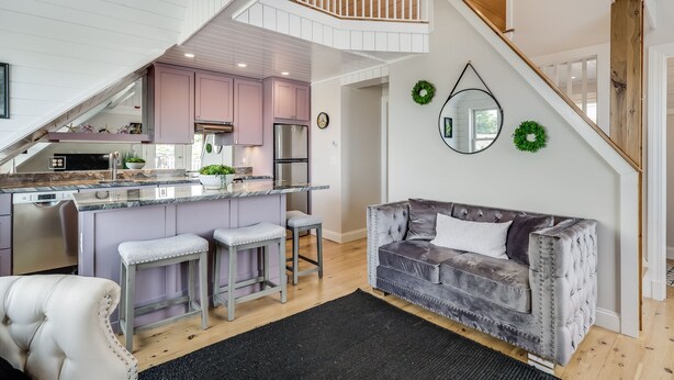 Een ruime woonkamer met een grijze, gewatteerde bank, een keuken met roze kastjes, en een trap naar een hoger niveau.