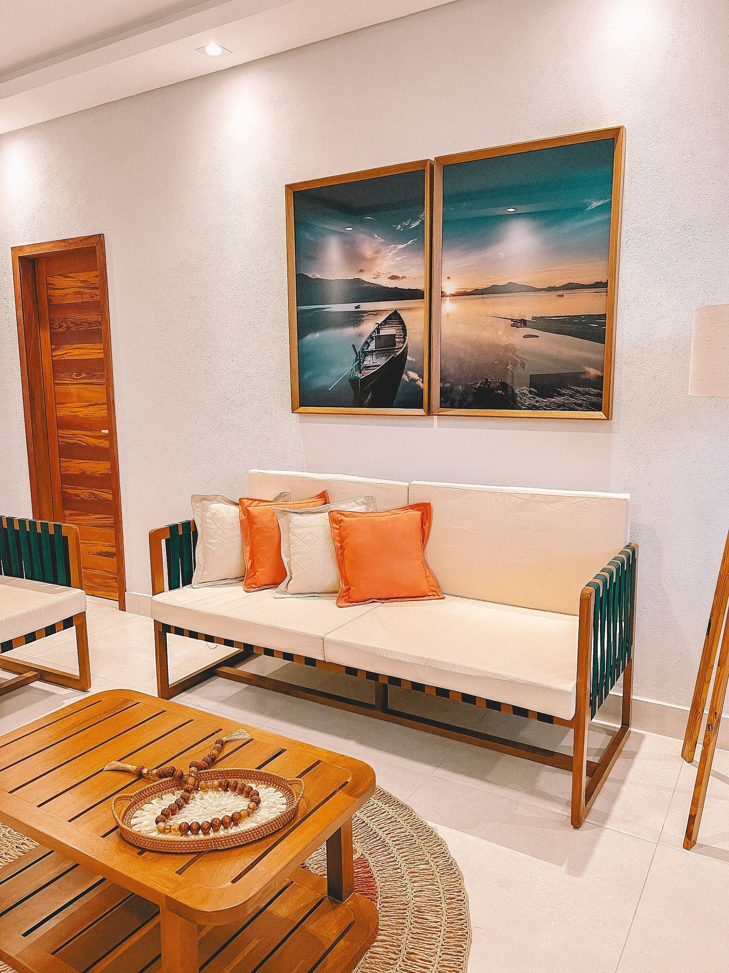 Sala de estar moderna com sofá branco, almofadas laranja, mesa de centro de madeira e duas pinturas emolduradas de paisagens na parede.