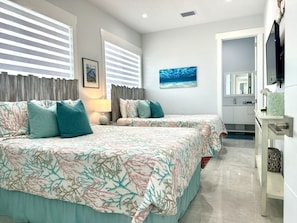Une chambre avec deux lits, une grande fenêtre avec des stores, un tableau représentant l’océan et une salle de bain visible à travers une porte ouverte.