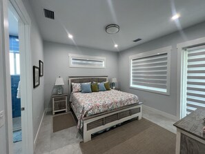A bedroom with a bed, bedside tables, a window with blinds, and a ceiling vent.