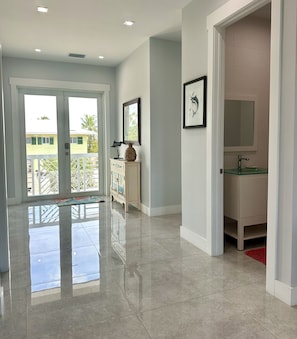 A spacious hallway with a polished floor, a white cabinet with decorative items, a framed picture, and a glass-top vanity with a red rug.