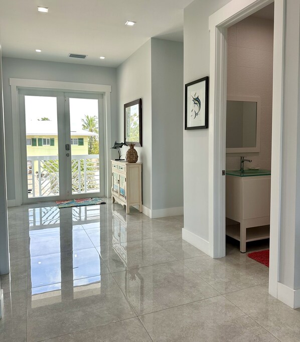 A spacious hallway with a polished floor, a white cabinet with decorative items, a framed picture, and a glass-top vanity with a red rug.