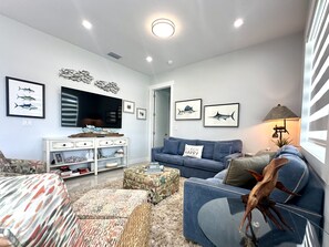 Un salon avec un canapé bleu, une console blanche, un téléviseur et des œuvres d’art représentant des poissons, encadrées et accrochées au mur.