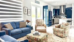 A modern living room with a blue sofa, a patterned ottoman, and a kitchen area with blue cabinets and a stainless steel refrigerator.