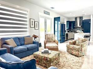 Una sala de estar moderna con un sofá azul, un taburete estampado y una cocina con armarios azules y un refrigerador de acero inoxidable.