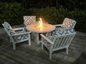 A round outdoor table with a built-in fire feature surrounded by four white chairs with patterned cushions.