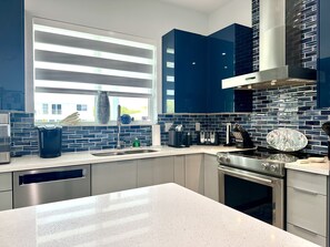 Une cuisine moderne dotée d’un grand îlot central, d’appareils électroménagers en acier inoxydable et offrant une vue sur l’extérieur grâce à une fenêtre munie de stores horizontaux.