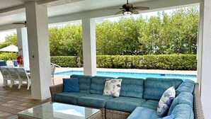 A covered outdoor seating area with a pool, wicker furniture, and a ceiling fan.