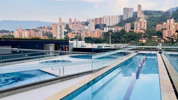 A rooftop swimming pool with clear blue water, surrounded by glass railings, and overlooking a cityscape with high-rise buildings and hills in the background. - 26th Floor A/C, Pool & Coworking (Medellín)