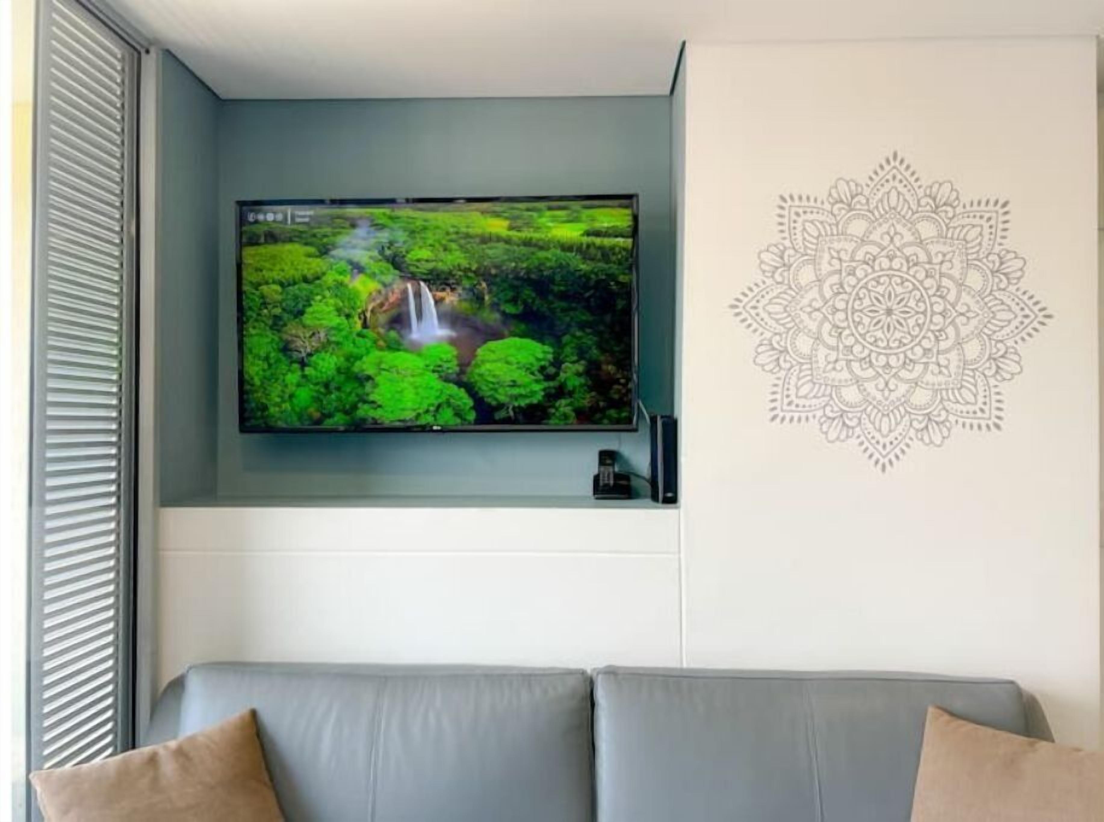 Ein Flachbildfernseher an der Wand über einer grauen Couch, mit einem dekorativen Mandalas auf der rechten Seite.