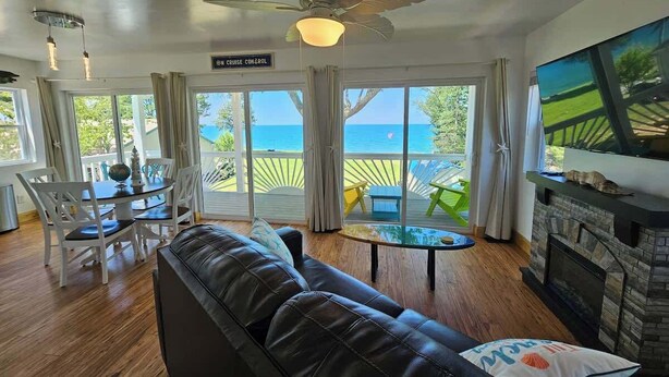A living room with a leather sofa, a dining table with chairs, a TV, a fireplace, and large windows overlooking the ocean. - Tree-Top Lake-Front Cottage (Madison)