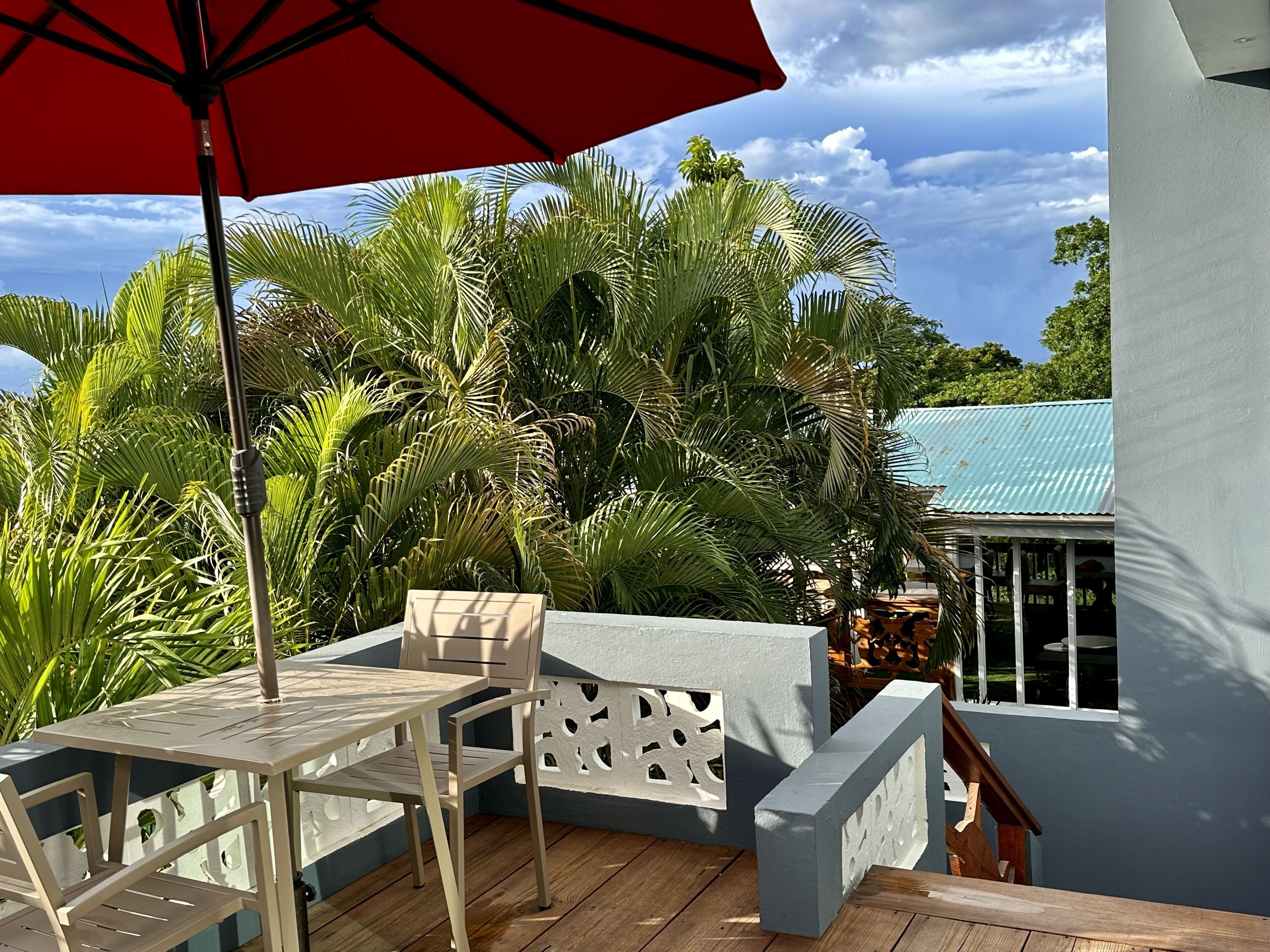 A balcony with a table and chairs, surrounded by lush palm trees and a blue-roofed building.