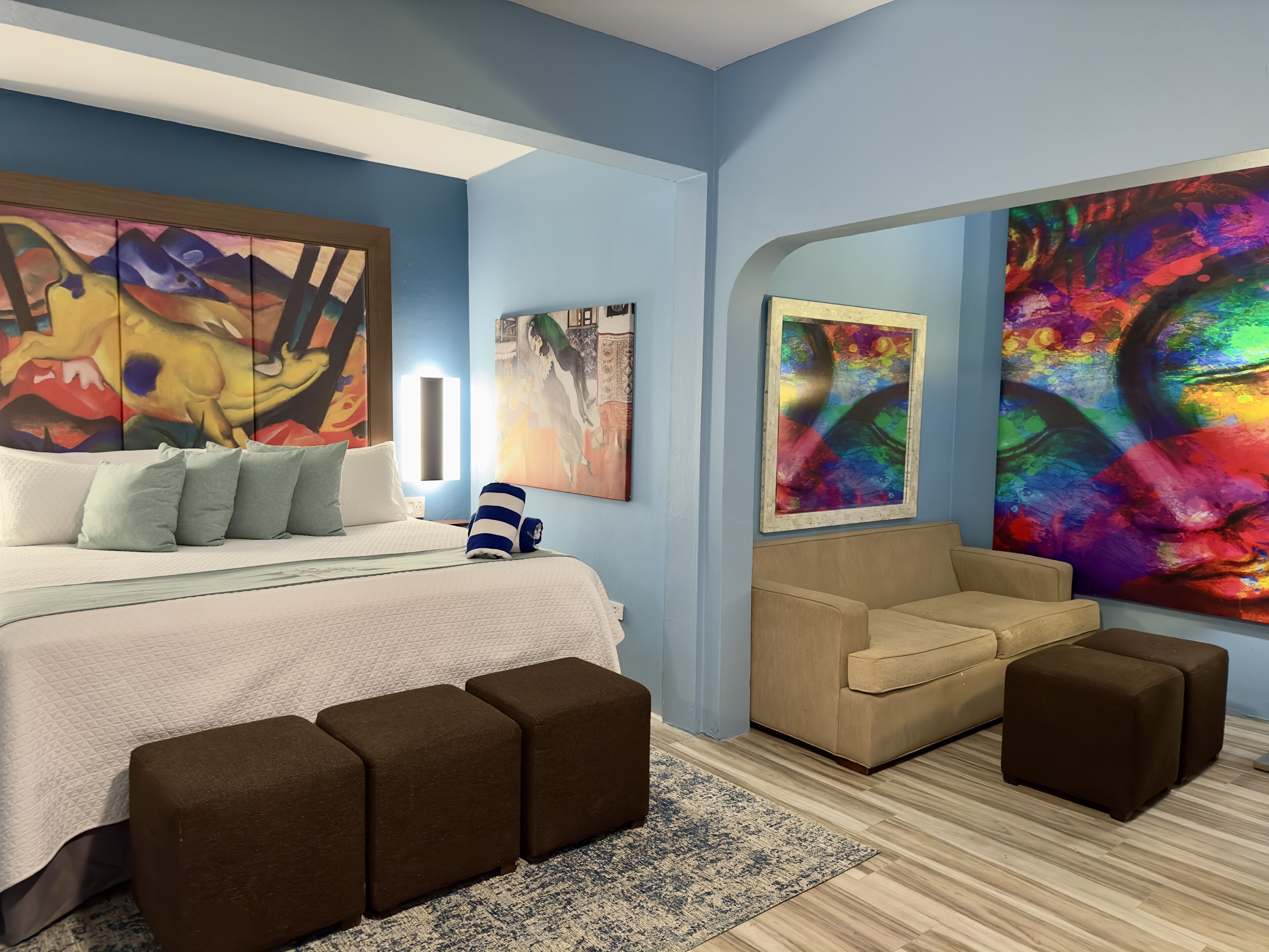 Quarto moderno com uma cama grande, um sofá e dois banquinhos. Há quadros abstratos nas paredes e uma janela com vista para a cidade.