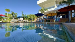 Piscina cristalina com espreguiçadeiras e guarda-sóis, cercada por palmeiras e um edifício com varanda.